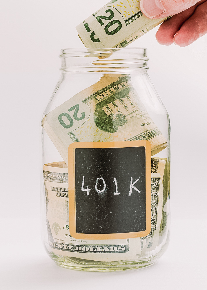 A hand depositing money into a glass jar labeled "401K" filled with US dollars.